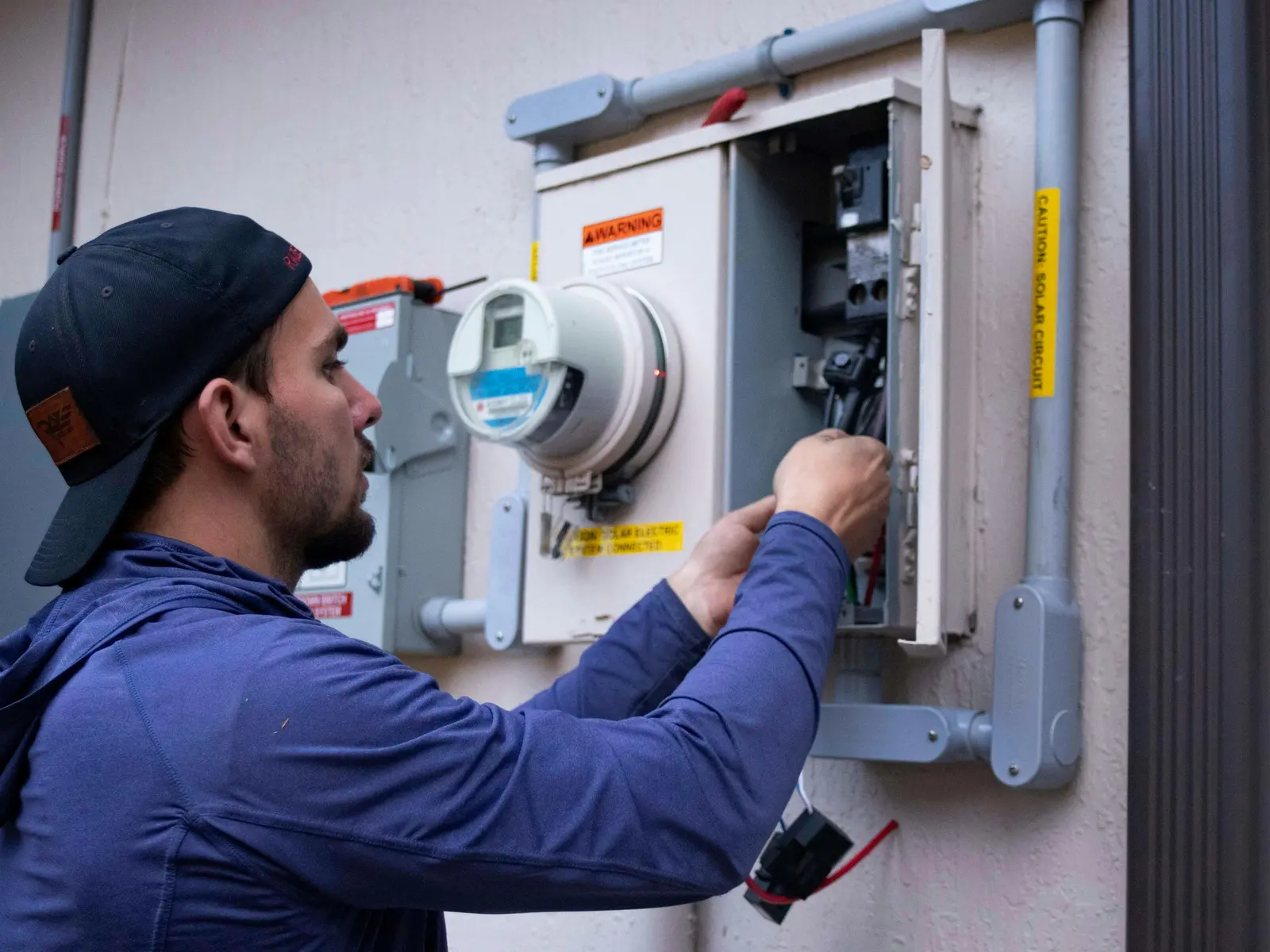 An electrician works inside an open meter and breaker panel on the outside wall of a house